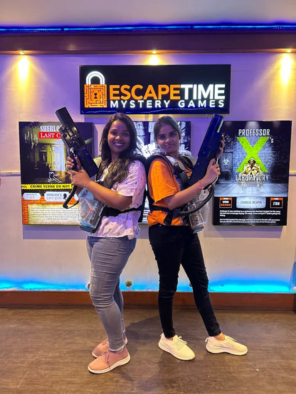 Two young women in a laser tag arena, holding phasers and wearing vests, stand in front of escape room posters.