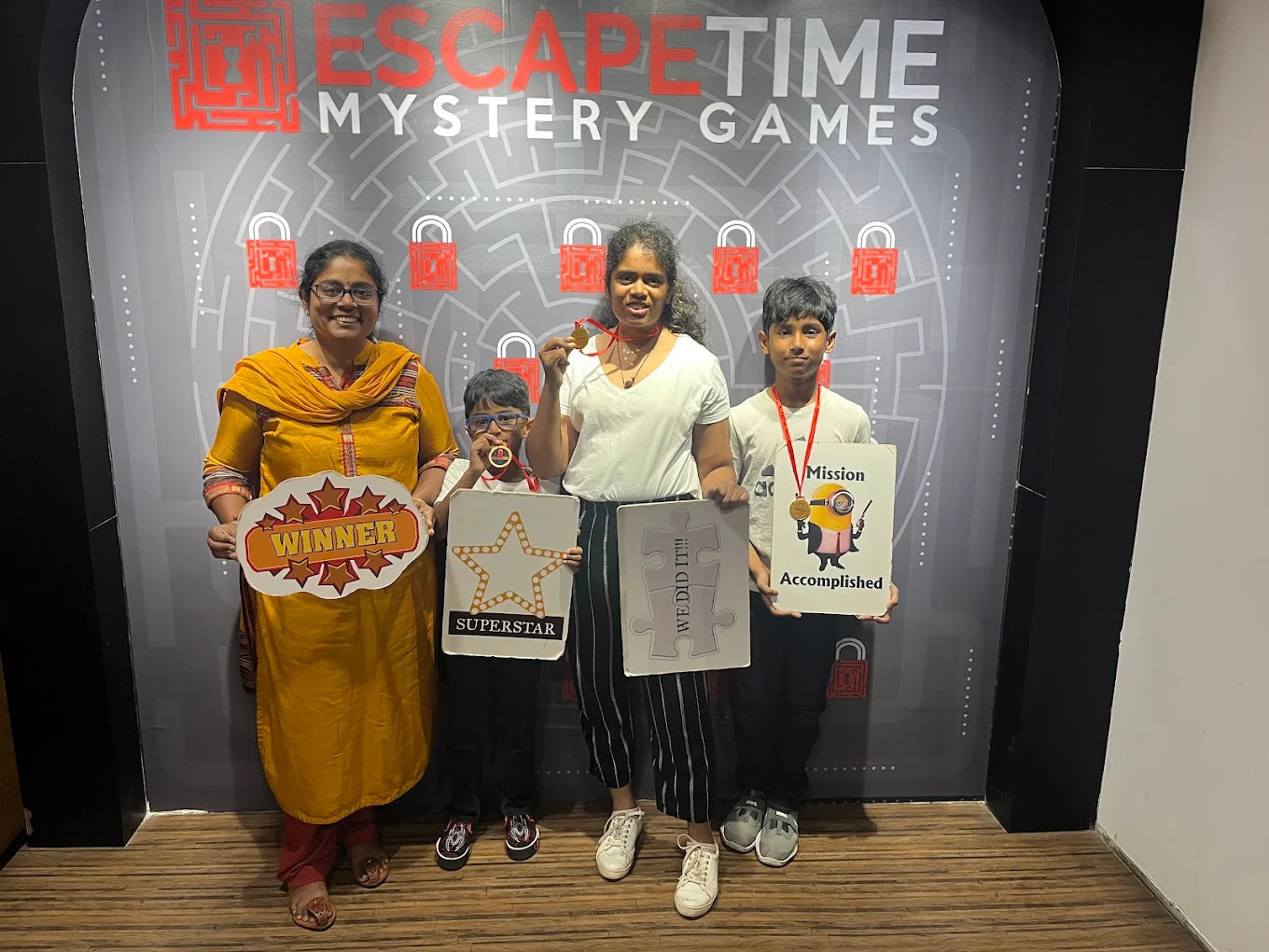 A family celebrates winning an escape room, holding signs and medals in front of a themed wall.