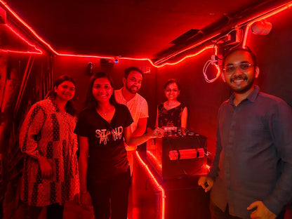 Four young adults pose in a room lit by red neon lights, with one person standing near DJ equipment.