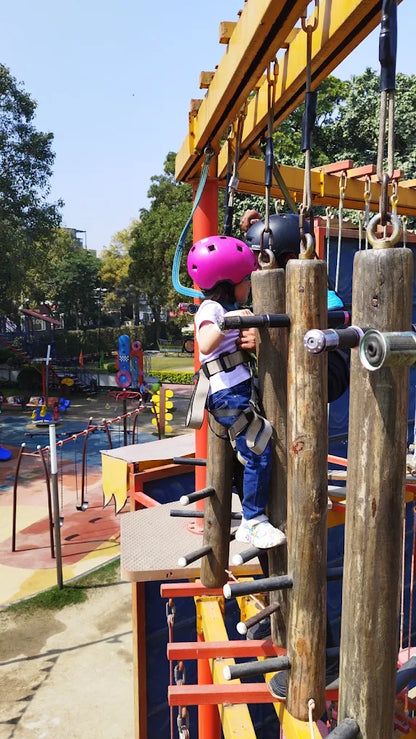 A young child in a pink helmet climbs a wooden obstacle course at a playground.