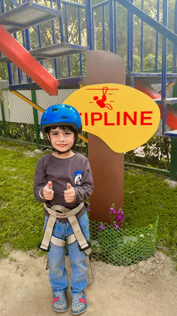 A young child in a helmet and harness gives a thumbs-up in front of a zipline sign.