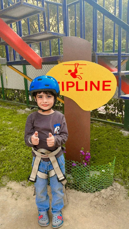A young child in a helmet and harness gives a thumbs-up in front of a zipline sign.