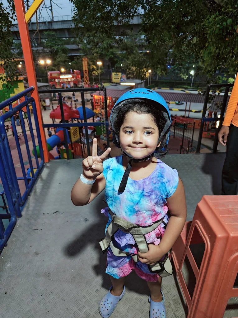 A young child in a helmet and harness poses for a photo at an outdoor adventure park.