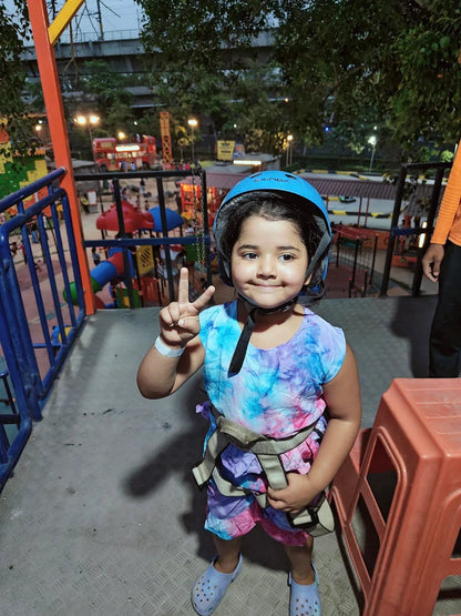 A young child in a helmet and harness poses for a photo at an outdoor adventure park.