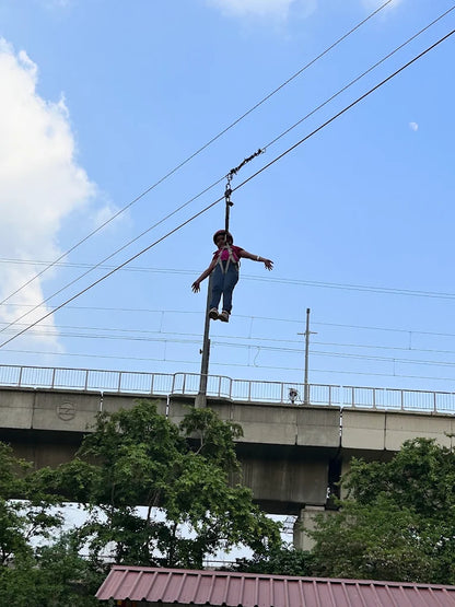A person in a harness ziplines across a clear sky, with a bridge and trees below.