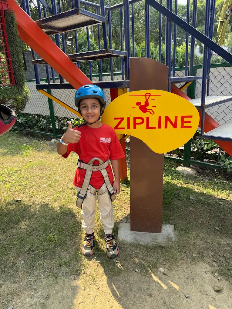 A young boy in a helmet and harness gives a thumbs-up in front of a zipline sign at an adventure park.