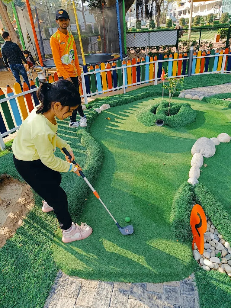 A young girl plays mini golf on a bright green course, with a man in an orange shirt nearby.