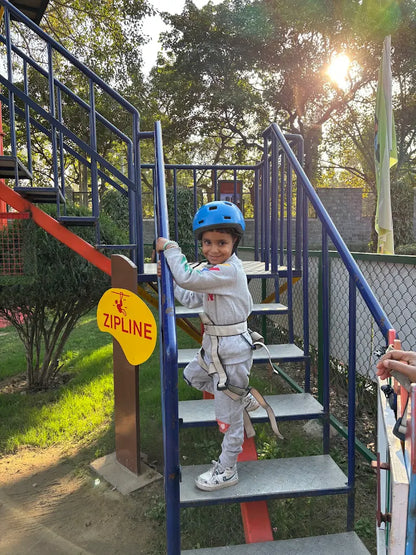 A young child in a helmet and harness climbs stairs on a zipline platform.