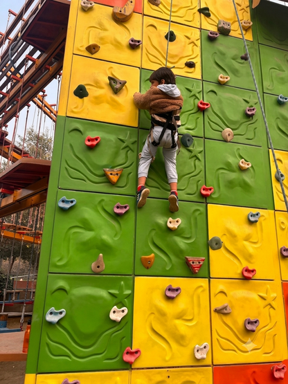 A child in a harness climbs a colorful outdoor rock wall.