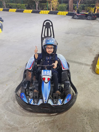 A young child in a helmet gives a thumbs-up while sitting in a go-kart at an outdoor track.