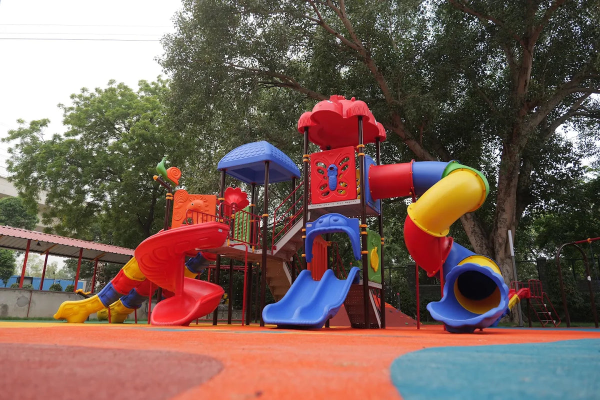 A colorful playground with slides and climbing structures for children, set outdoors under large trees.