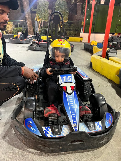 A young child in a go-kart helmet sits in a go-kart, with an adult's hands on the steering wheel.