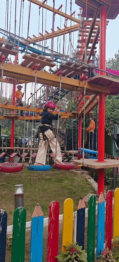 A young girl in a helmet navigates a ropes course, with another person visible in the background.