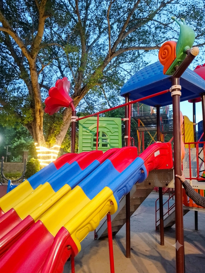 A colorful playground with slides and a snail decoration, set against a twilight sky and trees.