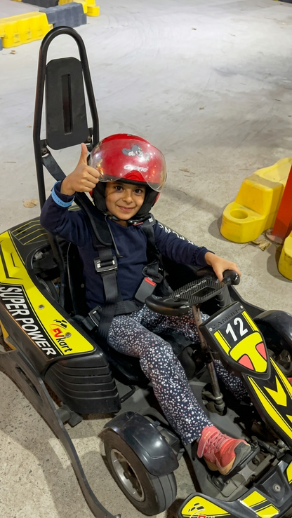 A young child in a red helmet gives a thumbs-up while sitting in a go-kart.