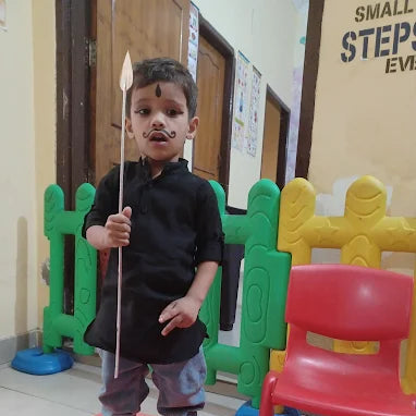 Child in a black shirt and gray pants holding a stick indoors with colorful furniture in the background.