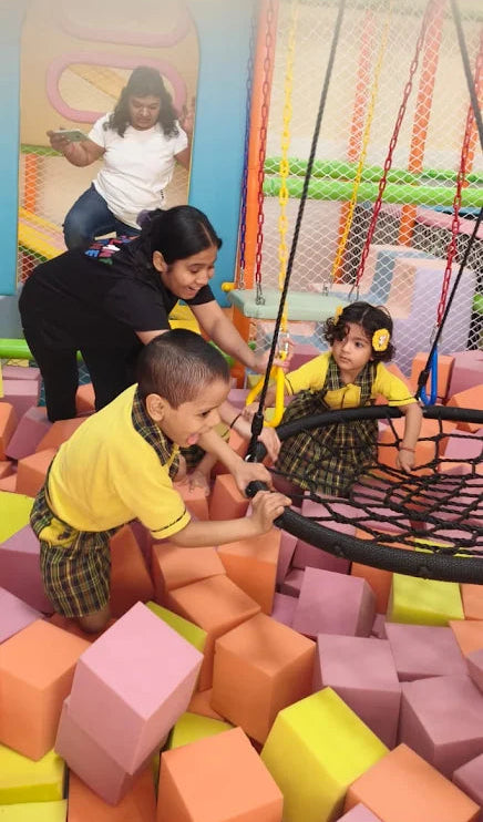 Young children playing on a large suspended net swing surrounded by soft foam blocks in an indoor play area