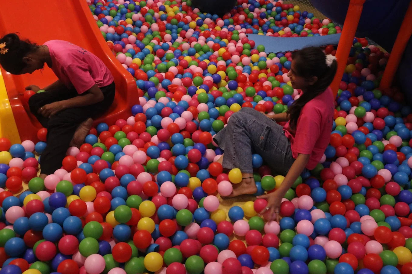Two children enjoying a large colorful ball pool with orange slides inside a spacious indoor play area