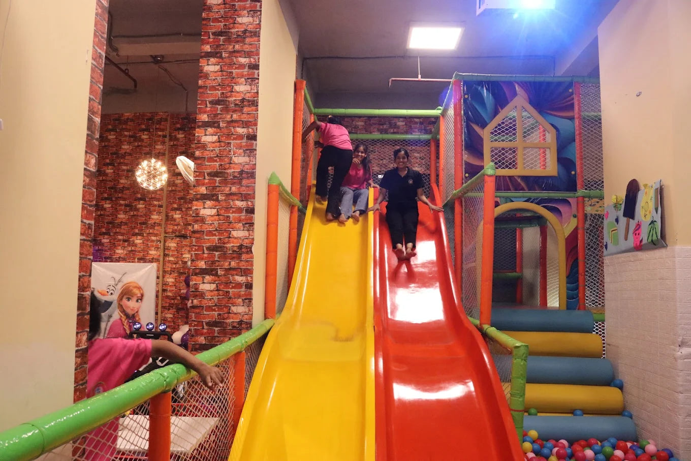Children sliding down bright yellow and red slides into a ball pit inside a colorful indoor play center