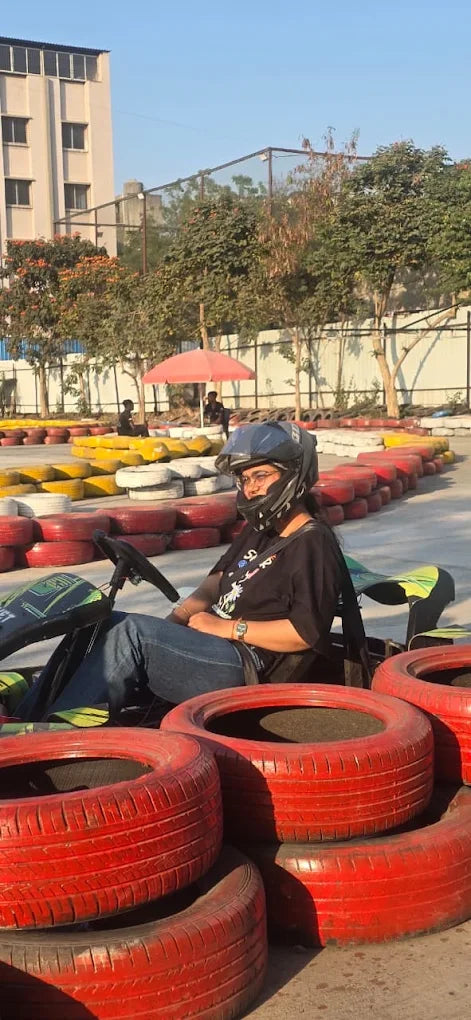 A young woman in a helmet sits in a go-kart at a track with red and yellow tire barriers.
