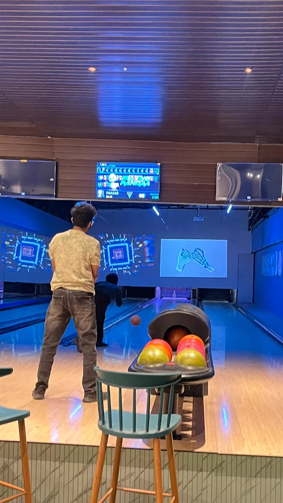 A young man bowls in a brightly lit bowling alley, with a bowling ball mid-air.