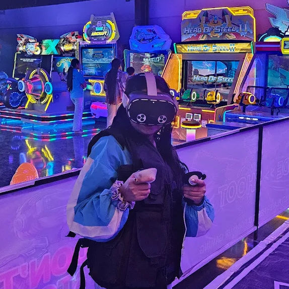 A young woman plays a VR game at an arcade, wearing a headset and holding controllers.