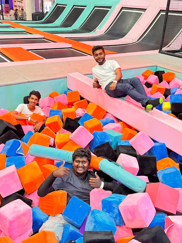 Two young men enjoy a trampoline park, one sitting on a pink beam and the other in a ball pit.