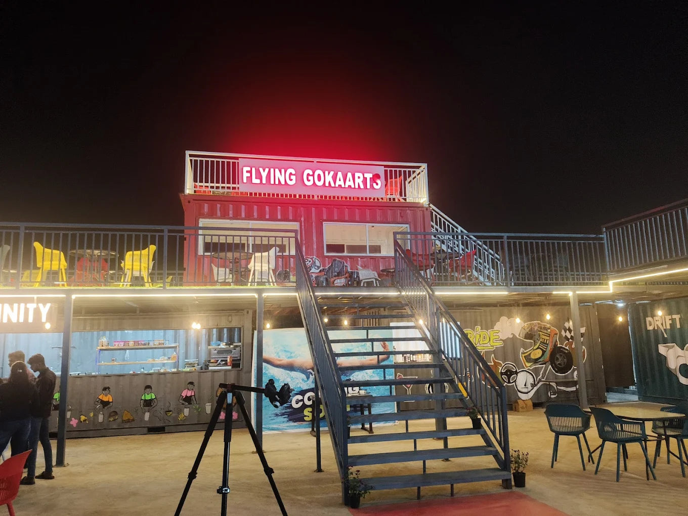 A multi-level outdoor entertainment venue at night, featuring shipping containers, stairs, and seating areas.