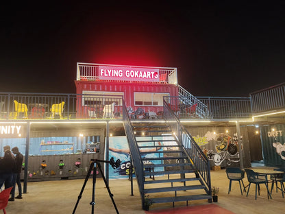 A multi-level outdoor entertainment venue at night, featuring shipping containers, stairs, and seating areas.