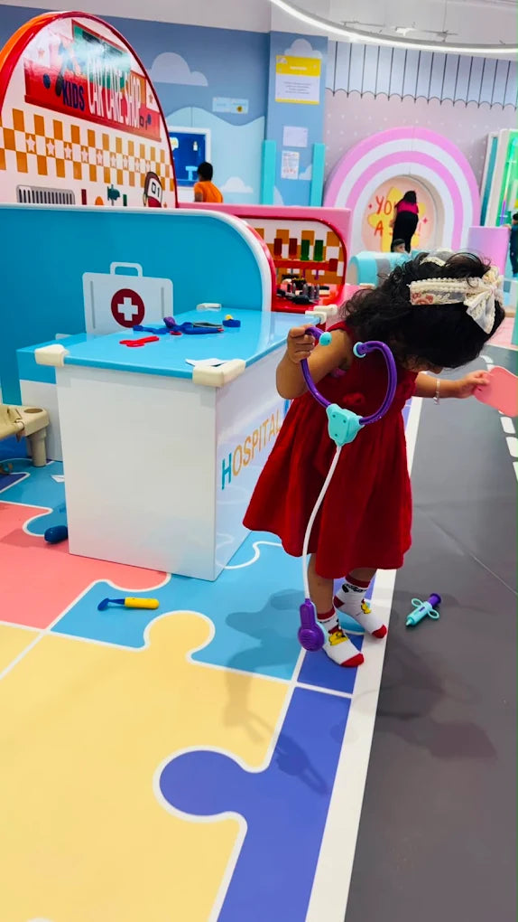 A young child in a red dress plays doctor with a toy stethoscope in a colorful hospital-themed play area.