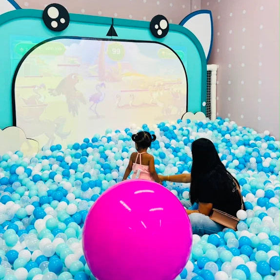 A young child and an adult play in a ball pit in front of a screen displaying a game.