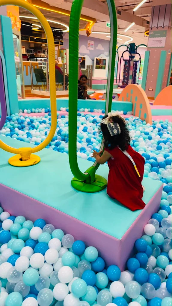 A young child in a red dress plays in a ball pit filled with blue and white balls. The child is reaching for a green tube in a colorful indoor playground.