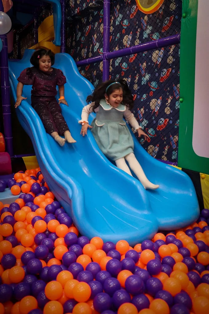 Two girls sliding down bright blue double slide into purple and orange ball pit at Fun Orbbit Kids Play Area Gujranwala Town Delhi