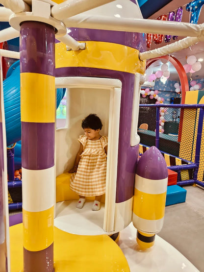 Young child in striped dress sitting inside colorful yellow and purple rocket play structure at Fun Orbbit Kids Play Area Gujranwala Town