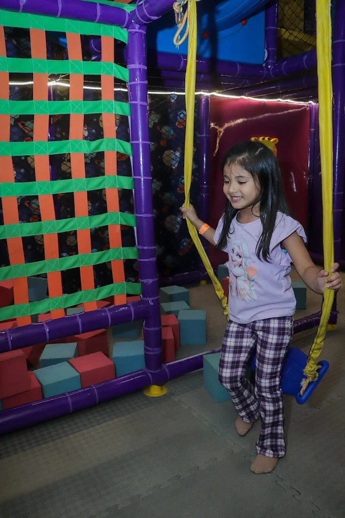 Smiling girl in purple shirt and plaid pants playing on yellow swing inside colorful indoor play area at Fun Orbbit Kids Play Area Gujranwala Town