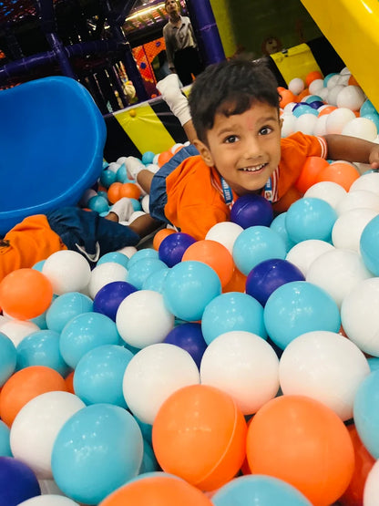 Boy in orange shirt smiling while lying in a colorful ball pit with blue slide in the background at Fun Orbbit Gujranwala Town