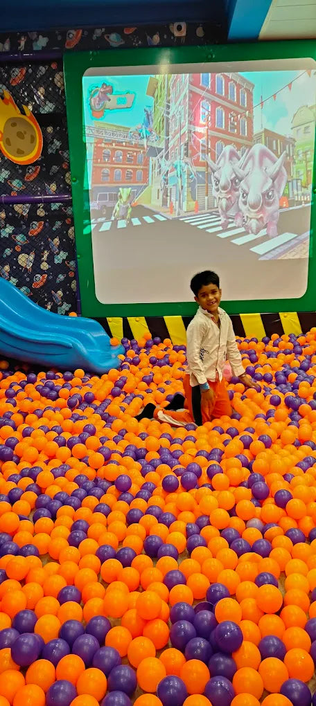 Child playing in a ball pit with colorful balls, watching a cartoon on a screen.