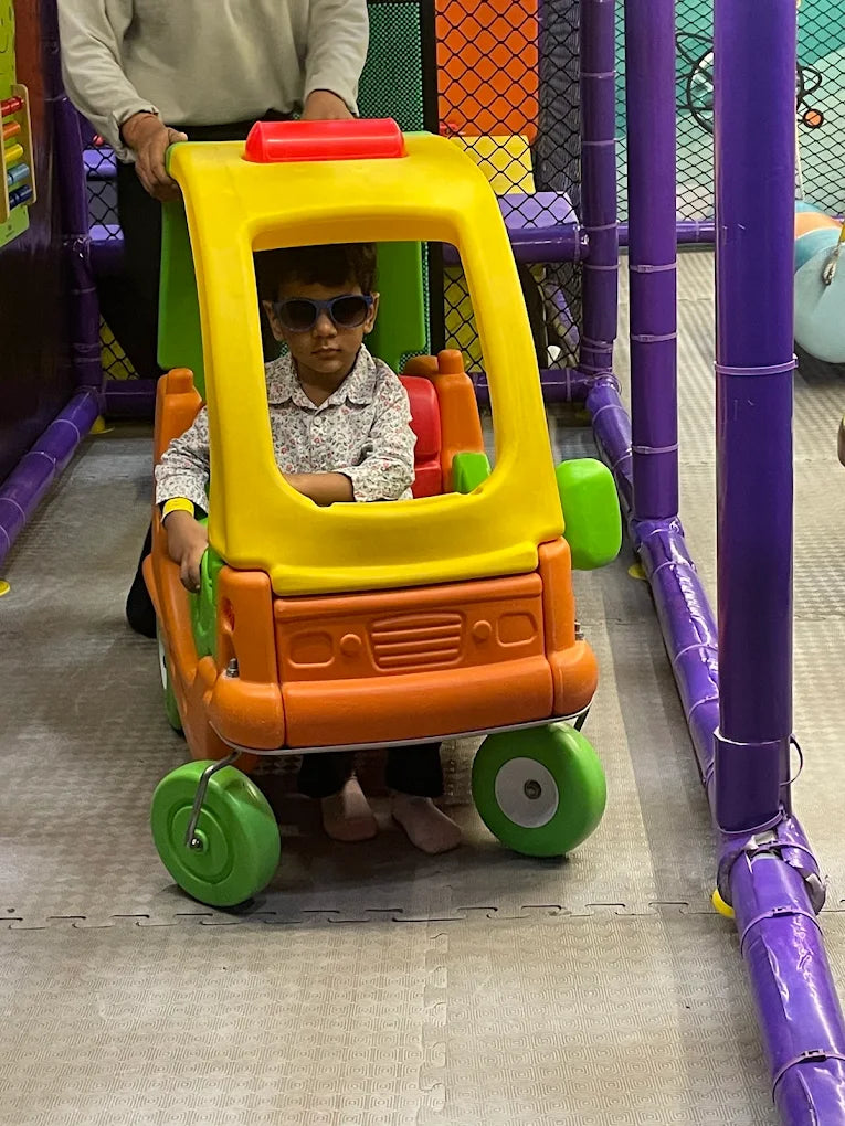 Young child wearing sunglasses sitting in bright orange and yellow toy car with adult nearby at Fun Orbbit Gujranwala Town Delhi