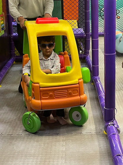 Young child wearing sunglasses sitting in bright orange and yellow toy car with adult nearby at Fun Orbbit Gujranwala Town Delhi