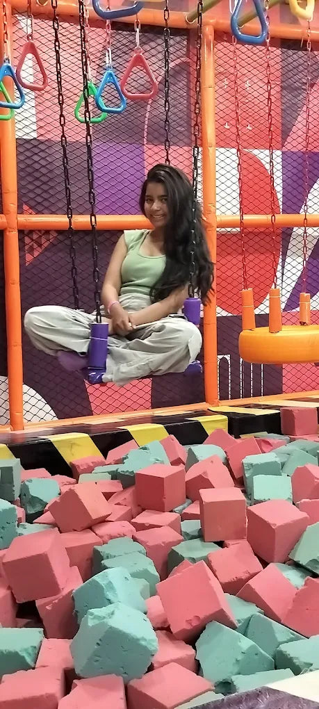 Teen girl sitting cross-legged on purple swinging seat above pink and teal foam pit at Fun Orbbit Kids Play Area Gujranwala Town Delhi