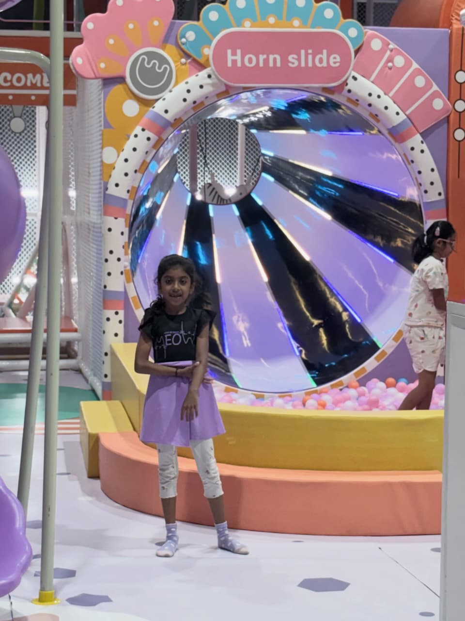A young girl stands in front of a colorful spinning tunnel at a playground. Another child is visible in the background.