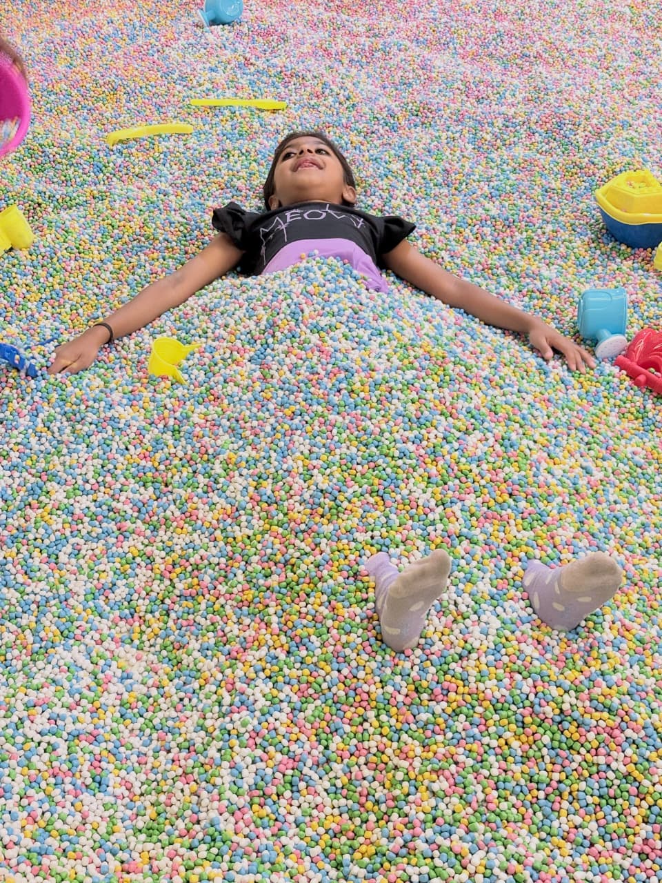 A young girl lies in a ball pit filled with colorful balls, with toys scattered around.