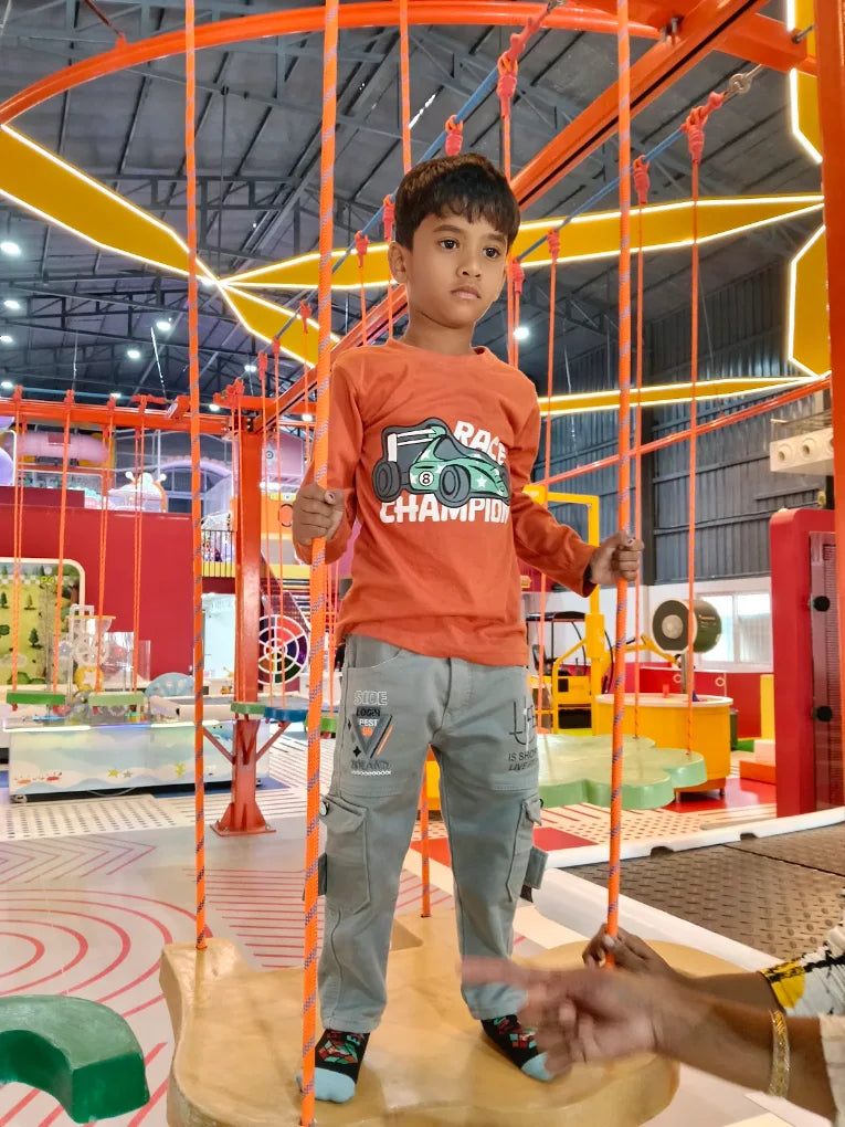 A young boy stands on a suspended platform in an indoor adventure park, holding onto orange ropes.