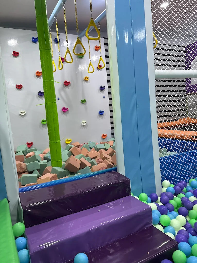 A colorful indoor playground with a climbing wall, foam pit, and ball pit for children.