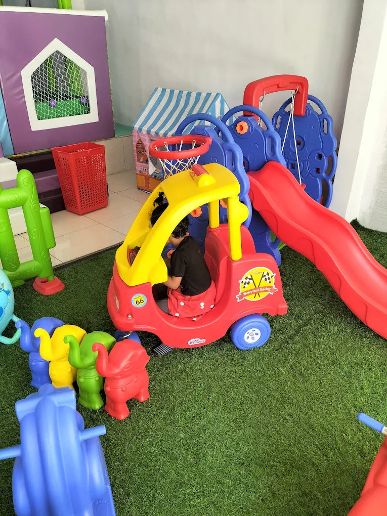 A child plays in a red toy car with a slide and basketball hoop in a playroom.