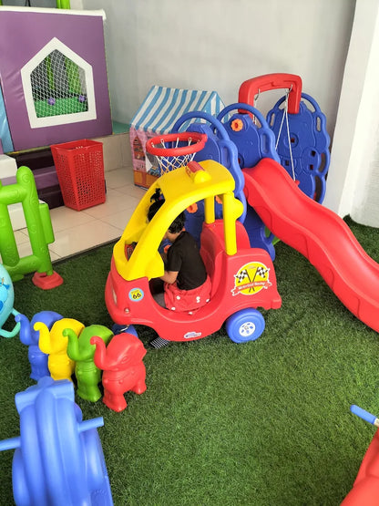A child plays in a red toy car with a slide and basketball hoop in a playroom.