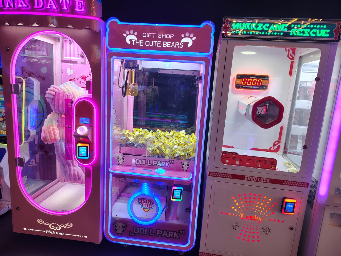 Three colorful arcade claw machines, one with a large teddy bear, one filled with yellow prizes, and one with a red and white design.