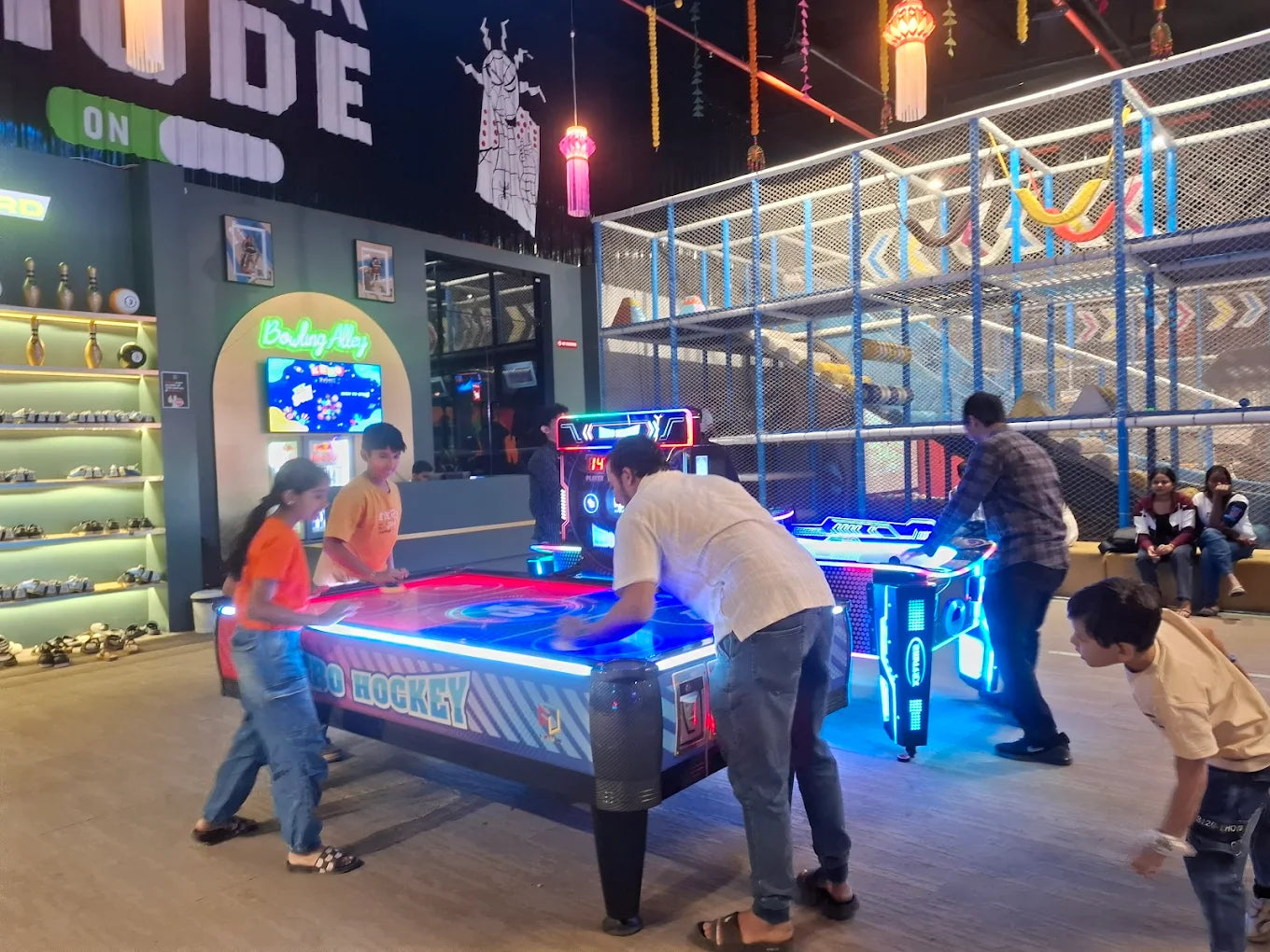 Children and adults play air hockey in a brightly lit arcade with a multi-level play structure in the background.