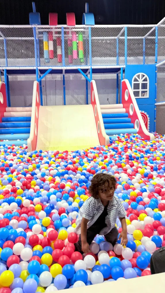 A young child plays in a ball pit with a large slide in the background.