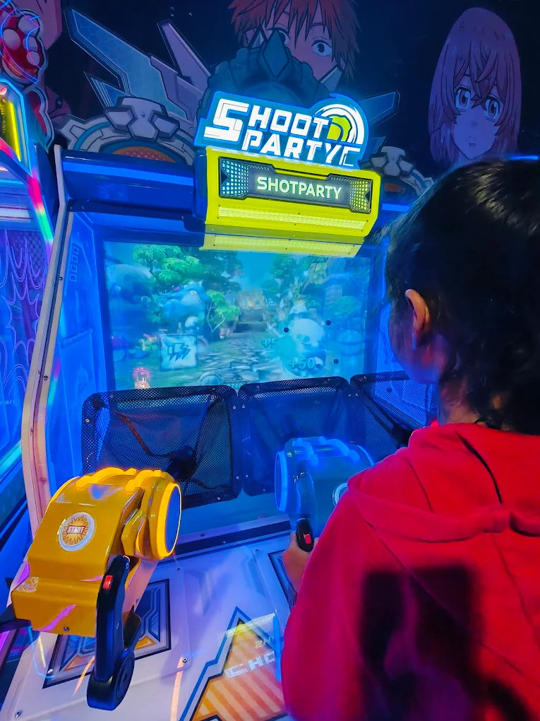 A child plays a shooting game at an arcade, holding a bright yellow toy gun. The game screen shows a colorful, animated scene.